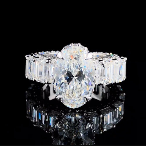 Front view of a white gold solitaire oval ring with emerald-cut stones on the side, displayed on a black background. A reflection of the ring on the bottom