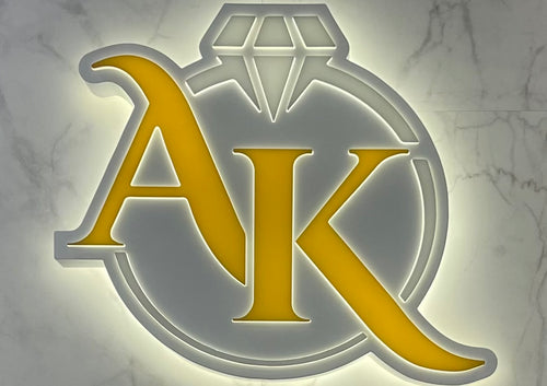 The letters 'AK' written in yellow inside a silver ring logo, displayed on a marble background.