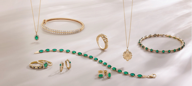 Emerald jewelry arranged on a cream background, including a bracelet, huggy earrings, emerald ring, bangle, necklace, diamond bangle, and gold ring, with 'Emerald and Diamond Jewelry Shop Now' text.