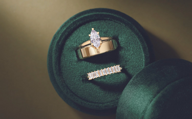 Three gold rings in a green velvet box: one marquise cut ring, a gold band, and an emerald cut band, with 'Jewelry for Everyone' written on top, displayed on a tan background.