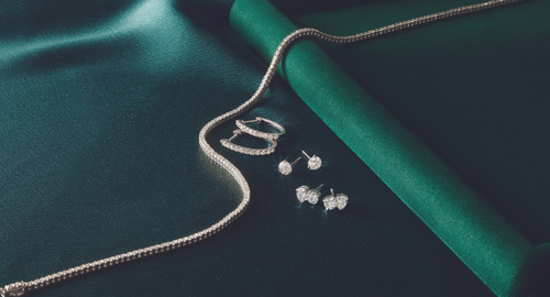 White gold tennis chain, three pairs of solitaire earrings, and diamond hoops, displayed on a dark green background.
