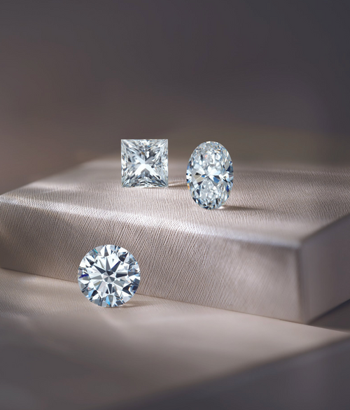 Three different shaped stones: round, oval, and princess cut, displayed on a tan display and riser.