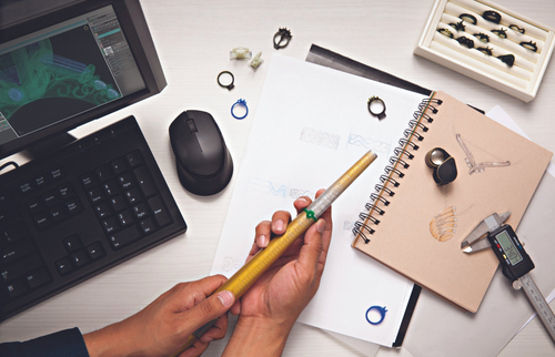 Computer with keyboard and mouse, gold mold ring on a ring mantel, millimeter gauge, notebooks, magnifying loop, and jewelry sketches on the desk