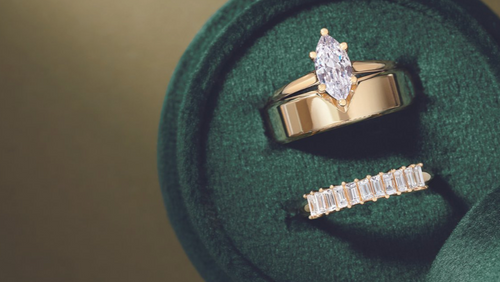 Three gold rings in a green velvet box: one marquise cut ring, a gold band, and an emerald cut band, with 'Jewelry for Everyone' written on top, displayed on a tan background.