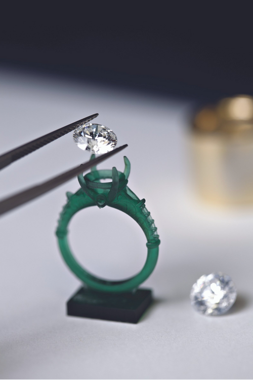 Diamond being set with tweezers on a green ring model, with a blurred stone next to it, displayed on a white background