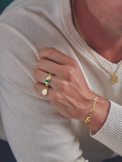 A man’s lower profile modeling jewelry including a gold bracelet, gold chain, and two rings, wearing a cream sweater on a teal background