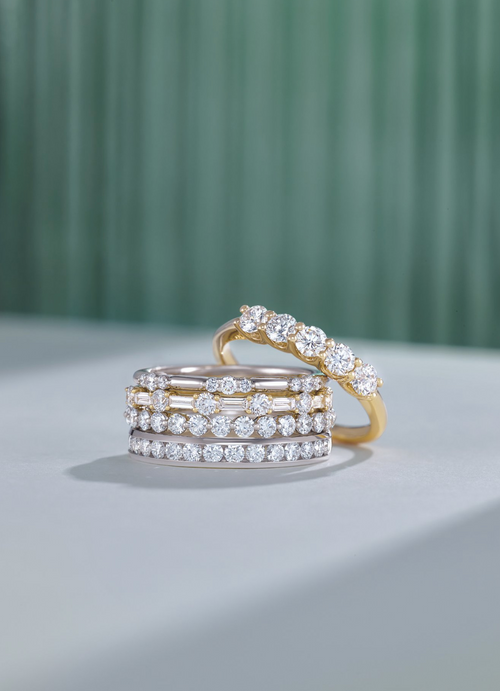 Four diamond bands rings stacked, with one leaning on the stack, displayed on a white surface with a teal background