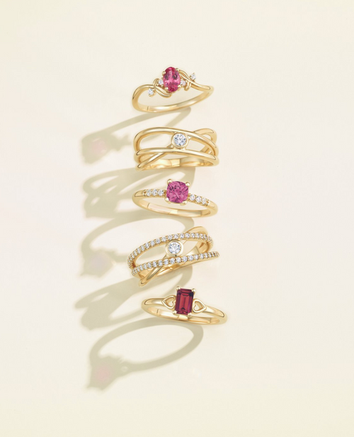 Five gold rings stacked: two with pink stones, one with a red tone, and two with white stones, displayed on a tan background.