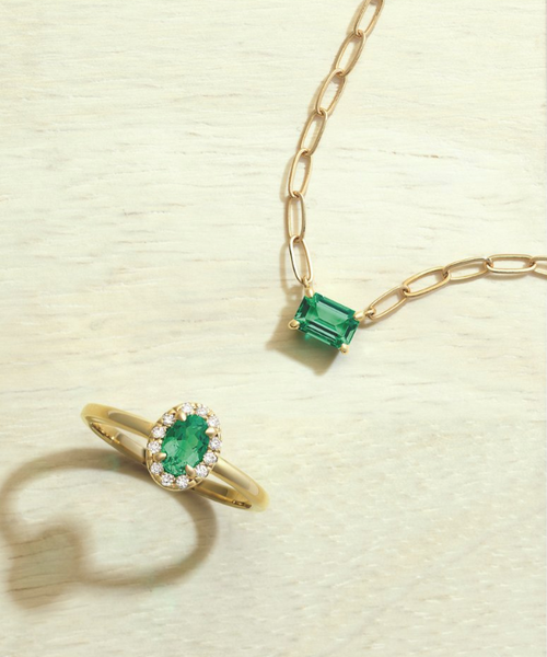 Emerald yellow gold ring with an emerald stone and a gold paperclip chain, displayed on a tan background